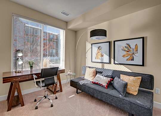 A bright home office here at Vista View featuring a large window with white blinds that fills the room with natural light, a wooden desk with a task chair and lamp, a modern gray sofa with patterned accent pillows, framed botanical wall art, a curved floor lamp, and soft carpeting creating a comfortable and functional workspace.