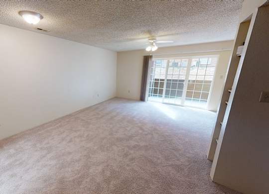 A spacious carpeted living room here at Windtree featuring soft neutral walls, plush carpeting, a ceiling fan with light fixture, ample open floor space, and large sliding glass doors that bring in natural light and provide access to a private outdoor area.