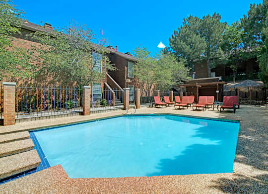 A sunny outdoor swimming pool here at Windtree featuring clear blue water, a textured pool deck, comfortable lounge chairs, shaded seating areas, mature trees, and surrounding brick apartment buildings that create a relaxed and inviting community setting.