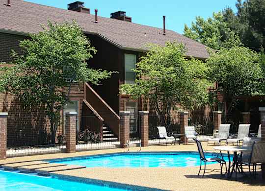 A sunny outdoor swimming pool here at Windtree featuring clear blue water, a curved pool edge with a raised lounging area, poolside tables and chairs, textured concrete decking, black metal fencing, mature trees, and brick apartment buildings creating a relaxed community atmosphere.