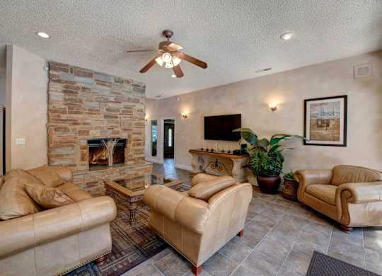 A warm community living room here at Windtree showcasing a stone fireplace as the focal point, plush leather seating arranged around a glass-top coffee table, tiled flooring with an area rug, soft wall sconces, a ceiling fan with lighting, and tasteful artwork creating a comfortable and inviting gathering space.