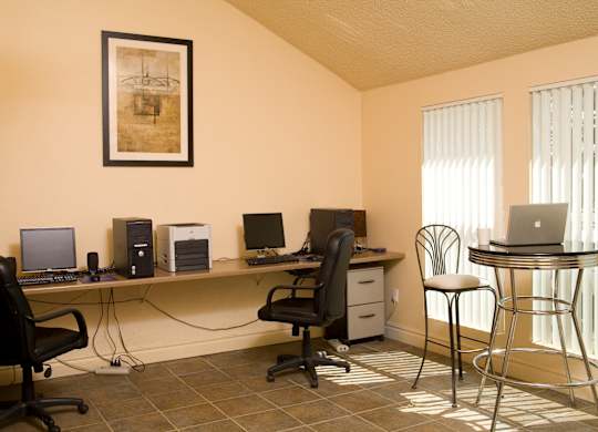 A bright community business center here at Windtree featuring a long shared desk with desktop computers and office chairs, a compact printer station, tiled flooring, a framed art piece on the wall, and tall windows with vertical blinds providing natural light alongside a small café-style table and seating.