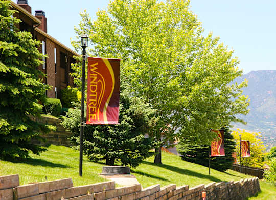 A landscaped hillside here at Windtree featuring manicured green grass, mature trees, and multiple branded Windtree banners displayed along a stone retaining wall, with apartment buildings partially visible to the left and scenic mountain views in the distance under bright, clear daylight.