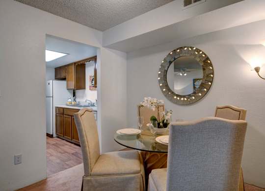 A warm and inviting dining area here at Windtree featuring a round glass-top dining table with upholstered chairs, soft carpet flooring, and a decorative wall mirror accented with subtle lighting, positioned just off the kitchen to create an open, comfortable space ideal for everyday meals and casual entertaining.