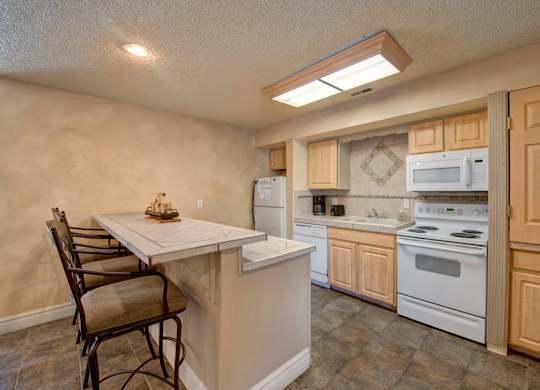 A cozy kitchen here at Windtree featuring light wood cabinetry, a peninsula-style counter with bar seating, white appliances, tiled flooring, and soft overhead lighting, creating a functional layout with ample counter space and an inviting area for casual dining and everyday cooking.