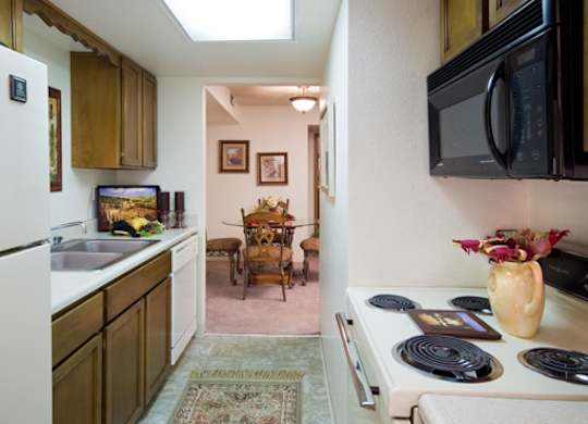 A warm kitchen here at Windtree featuring wood cabinetry, white appliances, a compact galley layout, and tiled flooring, with a view into the adjacent dining area that adds depth and flow while providing functional counter space and a practical design for everyday cooking and dining.