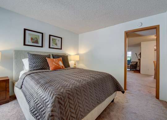 A cozy bedroom here at Windtree featuring plush carpeting, a neutral upholstered bed with textured gray bedding and accent pillows, warm bedside lighting, framed wall art above the headboard, and an open doorway leading into the adjoining living area, creating a comfortable and inviting private retreat.