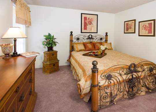 A traditional bedroom here at Windtree showcasing plush carpeting, an ornate metal and wood bed frame dressed in warm patterned bedding, matching decorative pillows, framed floral wall art above the headboard, a wooden dresser and nightstand, soft table lamp lighting, and classic furnishings that create a comfortable and welcoming atmosphere.
