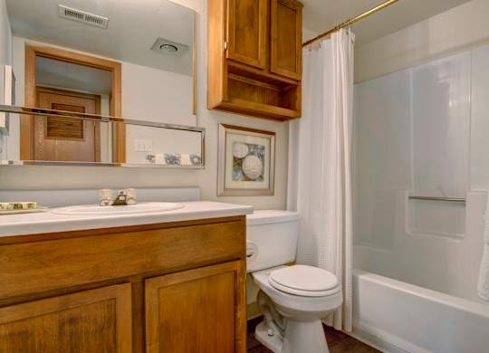 A clean bathroom here at Windtree featuring a wood-tone vanity with integrated sink, large wall mirror with shelving, white toilet, tub and shower combination with a textured surround and curtain, upper storage cabinet, framed wall art, neutral wall colors, and practical finishes that create a simple and functional space.