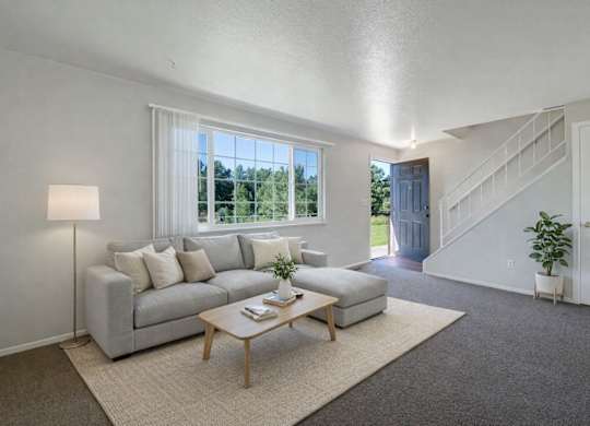 A bright modern living room here at Yorkshire Square featuring a light gray sectional sofa with neutral pillows, a simple wood coffee table on a soft area rug, and a large window bringing in natural light, with carpeted floors, an open stairway, and a welcoming entry that creates an airy, comfortable atmosphere.