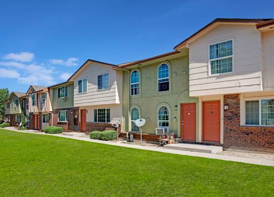 A bright exterior view here at Yorkshire Square featuring two-story townhome-style apartment buildings with neutral siding, brick accents, arched windows, private front entrances, and a well-maintained green lawn under a clear blue sky that highlights the welcoming residential setting.