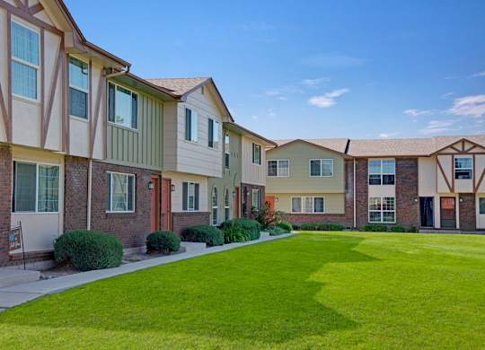 A charming exterior view here at Yorkshire Square showcasing two-story townhome-style apartment buildings with brick and light siding accents, varied architectural details, private entry doors, neatly trimmed shrubs, and a wide green lawn creating an inviting and well-kept residential atmosphere.