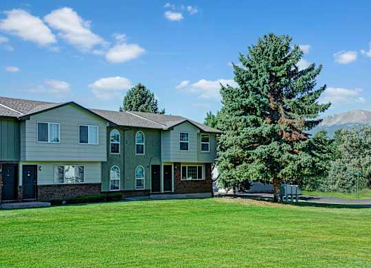 A spacious green courtyard here at Yorkshire Square featuring well-kept grass, mature evergreen trees, and a row of two-story apartment homes with neutral siding and brick accents, set beneath a bright blue sky with soft clouds and distant mountain views in the background.