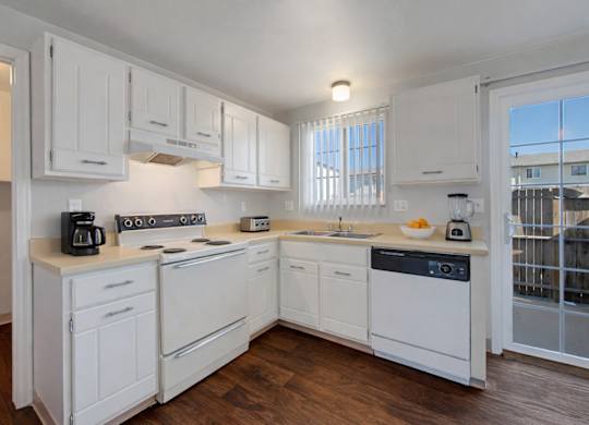 A bright, clean kitchen here at Yorkshire Square featuring white cabinetry, light countertops, and a matching appliance set, with a window over the sink for natural light, wood-style flooring, and a glass door leading outside that creates an open, functional, and inviting cooking space.