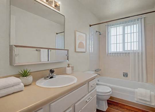 A bright, clean bathroom here at Yorkshire Square featuring a long vanity with a wide mirror and single sink, light cabinetry with ample storage, neutral countertops, a tub and shower combination with tiled surround, a window with vertical blinds for natural light, and warm flooring that adds contrast and comfort.