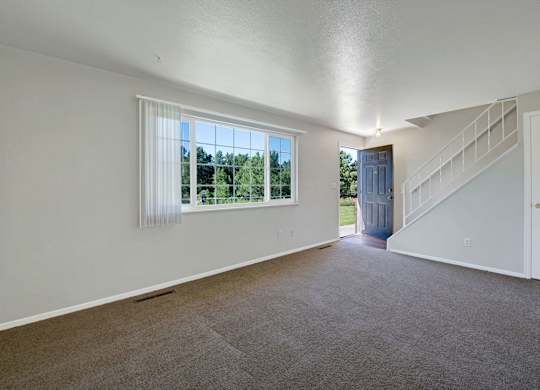 A bright, open living room here at Yorkshire Square featuring plush wall-to-wall carpeting, neutral painted walls, a large window with vertical blinds overlooking greenery, a private entry door, and a staircase with white railing that adds architectural interest and a sense of spacious flow to the layout.