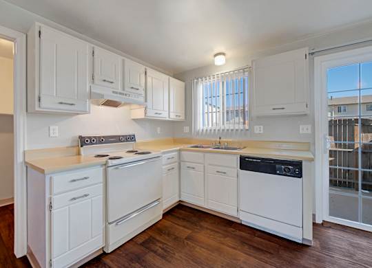 A bright, functional kitchen here at Yorkshire Square featuring white cabinetry with brushed hardware, neutral countertops, a freestanding range with overhead hood, a dishwasher, and a stainless sink beneath a window with vertical blinds, complemented by wood-style flooring and a sliding glass door that brings in natural light.