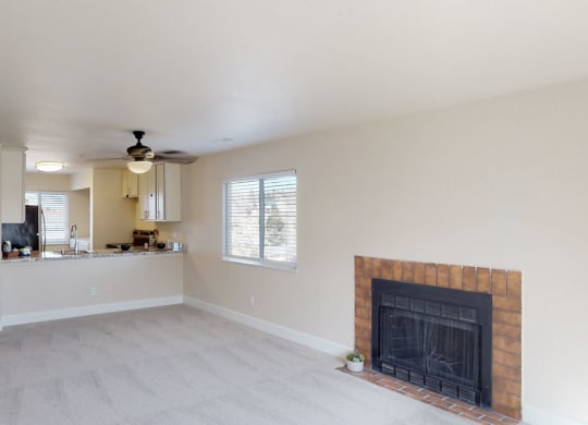Spacious living room here at Vista View Apartment Homes with beige carpet, a brick fireplace, and large windows. Open kitchen with white cabinets and a ceiling fan, creating a cozy atmosphere.