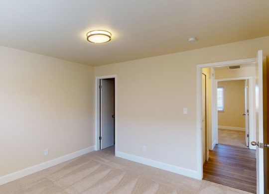 Small, empty room here at Vista View Apartment Homes with beige walls and carpet, illuminated by a ceiling light. Two open doors reveal a hallway and another room, creating a calm, neutral atmosphere.