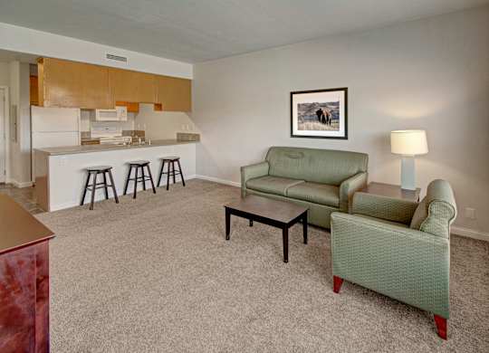 Cozy living room here at Confluence at the Harvest Hills Apartment Homes with green sofa and armchair, small coffee table, lamp, and framed bison picture. Open kitchen with three bar stools. Carpeted floor.