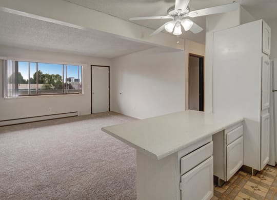 Spacious, empty living room here at The Continental Apartments with beige carpet, white walls, and large window. Kitchen with white cabinets and counter; ceiling fan present. Bright, airy feel.