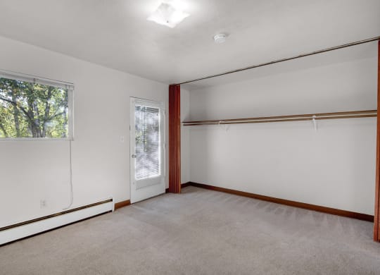 Living room here at Country Lane Apartments with beige carpet and white walls. A window with trees outside, a closet with a wooden rod, and a door with blinds are visible, creating a calm, spacious atmosphere.