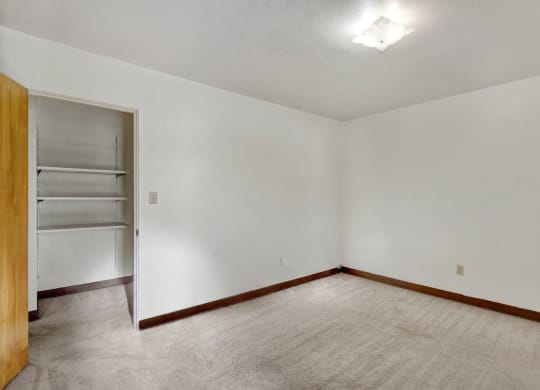 Empty bedroom here at Country Lane Apartments with beige carpet, white walls, and a wooden baseboard. An open closet with shelves is visible, and a ceiling light softly illuminates the space.