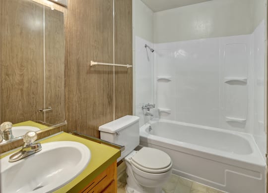 Small bathroom here at Country Lane Apartments with a white bathtub and shower, wood-paneled walls, and a yellow countertop with a sink. The atmosphere is simple and clean.