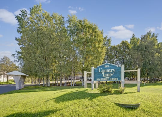 Sign reading "810 Country Lane Apartment Homes" stands on a lush, green lawn with a backdrop of tall trees under a clear blue sky, conveying tranquility.