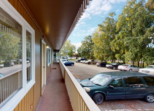 Outdoor hallway here at Country Lane Apartments with wooden siding, overlooking a parking lot. Several cars are parked next to lush green trees under a partly cloudy sky.