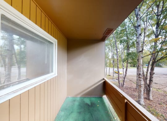 Small balcony here at Country Lane Apartments with green carpeted floor and brown wooden walls overlooks a parking area and trees. The setting is calm and outdoors.