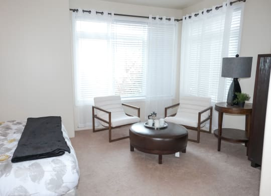 Bright bedroom here at Cheyenne Creek Apartment Homes with two white chairs, a brown leather ottoman, and a side table with a lamp. White curtains and soft lighting create a cozy, serene atmosphere.