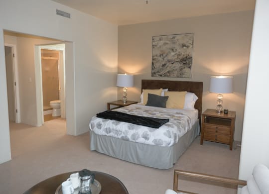 A cozy bedroom here at Cheyenne Creek Apartment Homes with a queen-sized bed, flanked by two wooden nightstands and lamps. Neutral tones, abstract art above, and an adjoining bathroom enhance the tranquil atmosphere.