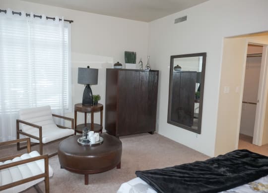Modern living room here at Cheyenne Creek Apartment Homes with beige walls, featuring two white cushioned chairs, a round brown coffee table with a tray, and a dark wooden cabinet. A tall lamp sits beside the cabinet, and a large wall mirror adds depth. The space has a calm, minimalist vibe.