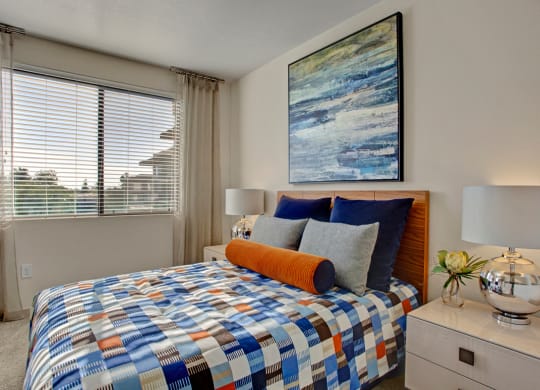 Modern bedroom here at Escape Apartments with a colorful patchwork bedspread, navy and gray pillows, and an orange bolster. Abstract wall art and a silver lamp add elegance.
