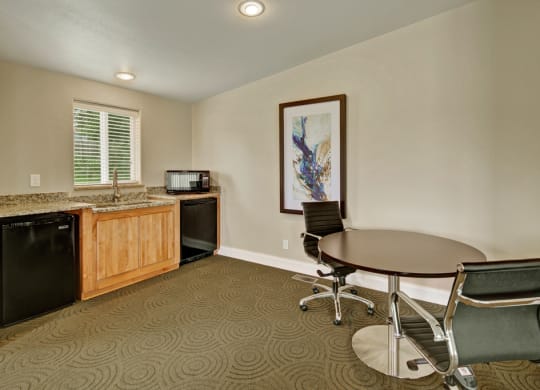 Minimalist office space here at Four Seasons Apartments with a round wooden table, two sleek black chairs, and a kitchenette with light wood cabinets. A colorful abstract painting adorns the beige wall.