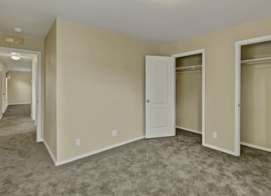 Bedroom here at Four Seasons Apartments with beige walls, open closet doors, and gray carpet. A hallway is visible leading to another lit room, creating a neutral and vacant atmosphere.