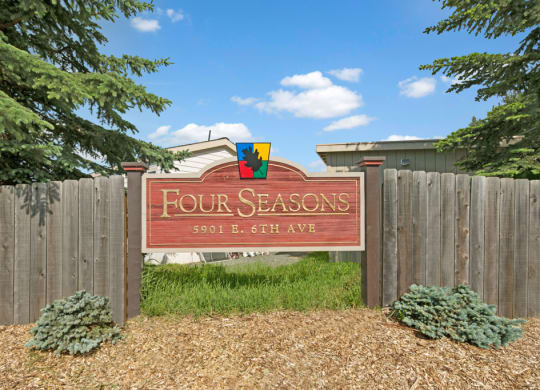 Wooden sign reading "Four Seasons, 5901 E. 6th Ave" surrounded by a wooden fence, green shrubs, and pine trees under a clear blue sky.