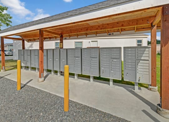 A covered outdoor mail station here at Four Seasons Apartments with multiple gray mailboxes lined up under a wooden shelter. Yellow bollards are in front on a gravel surface.