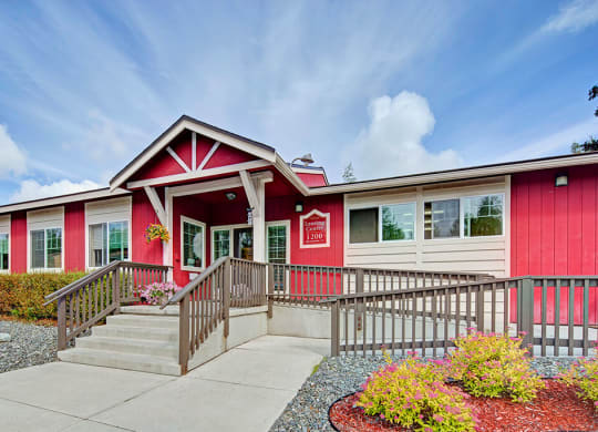 Front view of a red and white building here at The Glen Apartments, identified as a Leasing Center. The entrance features steps and a ramp, surrounded by colorful landscaping under a partly cloudy sky.