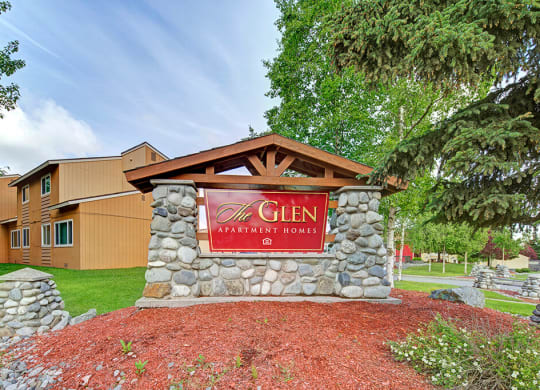Stone sign for "The Glen Apartment Homes" near a tan building, surrounded by lush greenery and pine trees, under a clear, blue sky. Calm atmosphere.
