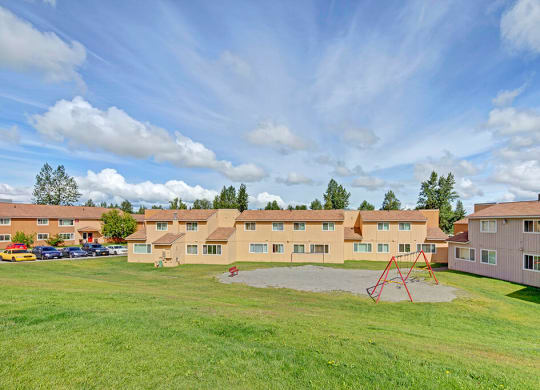 The Glen Apartment complex under a bright blue sky with clouds. Yellow buildings have brown roofs and are surrounded by green grass. A red swing set is visible.