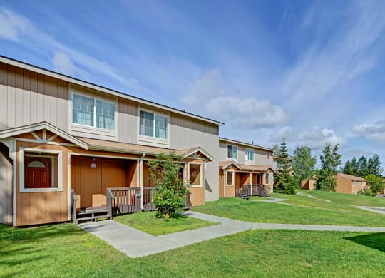 Two-story beige townhouses here at The Glen Apartments with porches, surrounded by green lawns and trees on a sunny day. The atmosphere is peaceful and suburban.