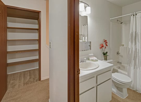 A clean bathroom here at The Glen Apartments with white fixtures, including a sink, toilet, and bathtub with a patterned curtain. A small red plant decorates the vanity. On the left, an empty closet with wooden shelves is visible, creating a tidy and minimalistic atmosphere.