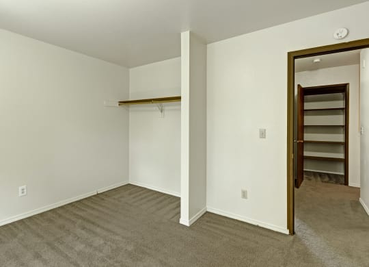 Empty closet here at The Glen Apartments with beige carpet and white walls, featuring a single wooden rod. An open door on the right leads to a room with shelving. Minimalist and clean.