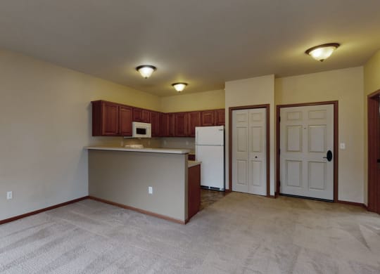 Interior of an empty apartment here at Grand River Estates Apartment Homes featuring a carpeted living area and a kitchen. The kitchen includes wooden cabinets, a white fridge, and a microwave. The walls are beige, and three ceiling lights brighten the space, creating a warm, inviting atmosphere.