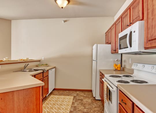 Warm kitchen here at Grand River Estates Apartment Homes with wood cabinets, white appliances, and beige countertops. A jar of oranges and neatly arranged dishware add a cozy, inviting feel.