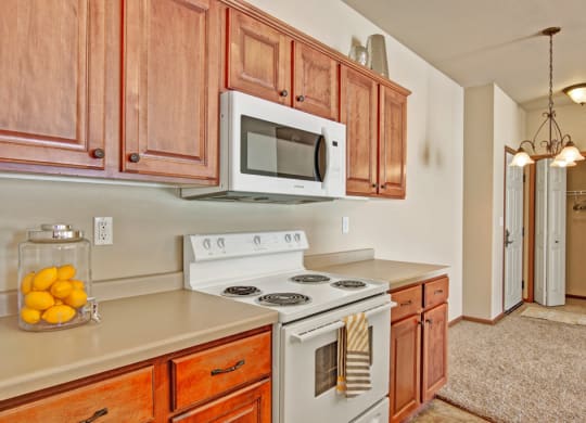 A cozy kitchen here at Grand River Estates Apartment Homes with wooden cabinets, a white stove, and a microwave. Lemons in a jar add a bright touch. Warm lighting sets an inviting tone.