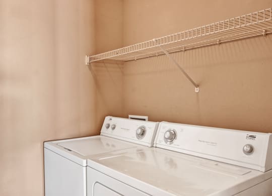A bright laundry room here at Grand River Estates Apartment Homes with a white washer and dryer side by side against a beige wall. An empty white wire shelf hangs above them.