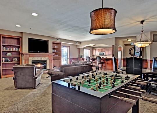 Cozy recreation room here at Grand River Estates Apartment Homes with a foosball table, armchairs by a fireplace, bookshelves, large TV, and bar area. Warm lighting and relaxed atmosphere.